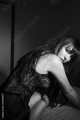 Black and white boudoir portrait of a caucasian woman wearing a black lace body suit. The photos have a film noir feel and have a sensual vibe to them. The lady has long dark hair with bangs.` © Angela