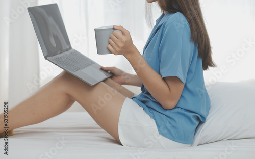 Young Asian woman with beautiful face, long hair, blue shirt lying on bed in white bedroom at home with cup of coffee and video call with laptop talking to relatives on vacation. holiday concept.
