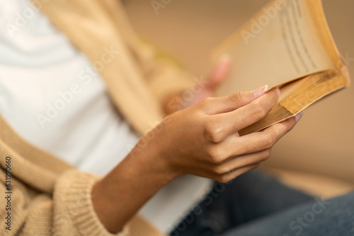 Close up female hands holding book turning the page. Young woman sitting on cozey couch sofa reading book in living room with warm light at night. People female stay home part time relax at home.