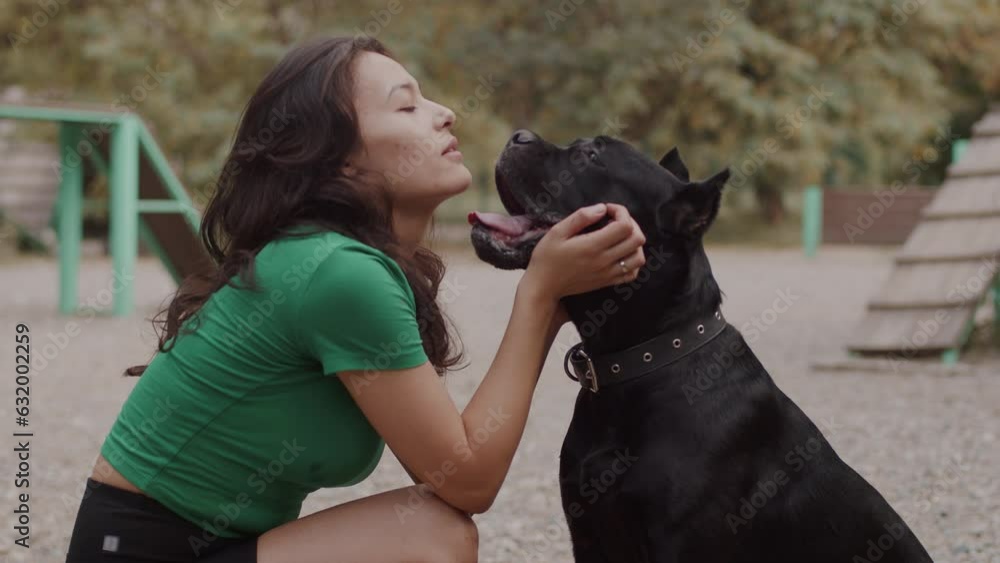 Young asian woman squatting at the outdoor dog training ground, patting her big black dog and talking sweetly with it. Concept of proper pet care and handling