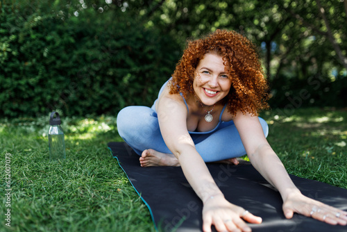 Portrait of happy pretty ginger head girl training outside in green area, sitting on mat with crossed legs, reaching hands out bending forward, smiling at camera. Yoga practice outdoor