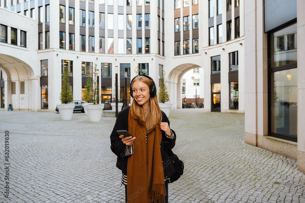 Fototapeta premium Smiling woman listening music with headphones and smartphone while standing outdoors