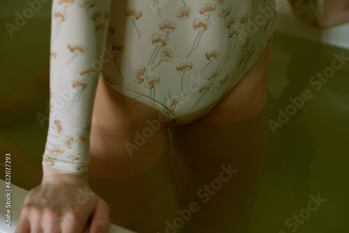 Close-up shot of slim girl in bodysuit with floral pattern standing in bath full of creepily green water, holding to its sides and getting ready to sink  photo