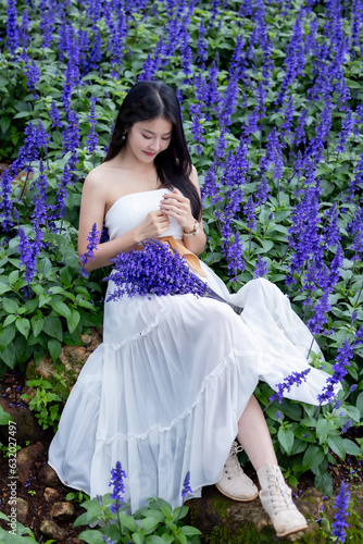 Asian woman in white dress sitting amidst in flowers field. Beautiful girl holding bouquet flowers in hands. Happy woman enjoying in purple flower field and nature sea of fog. Embrace nature,wind.