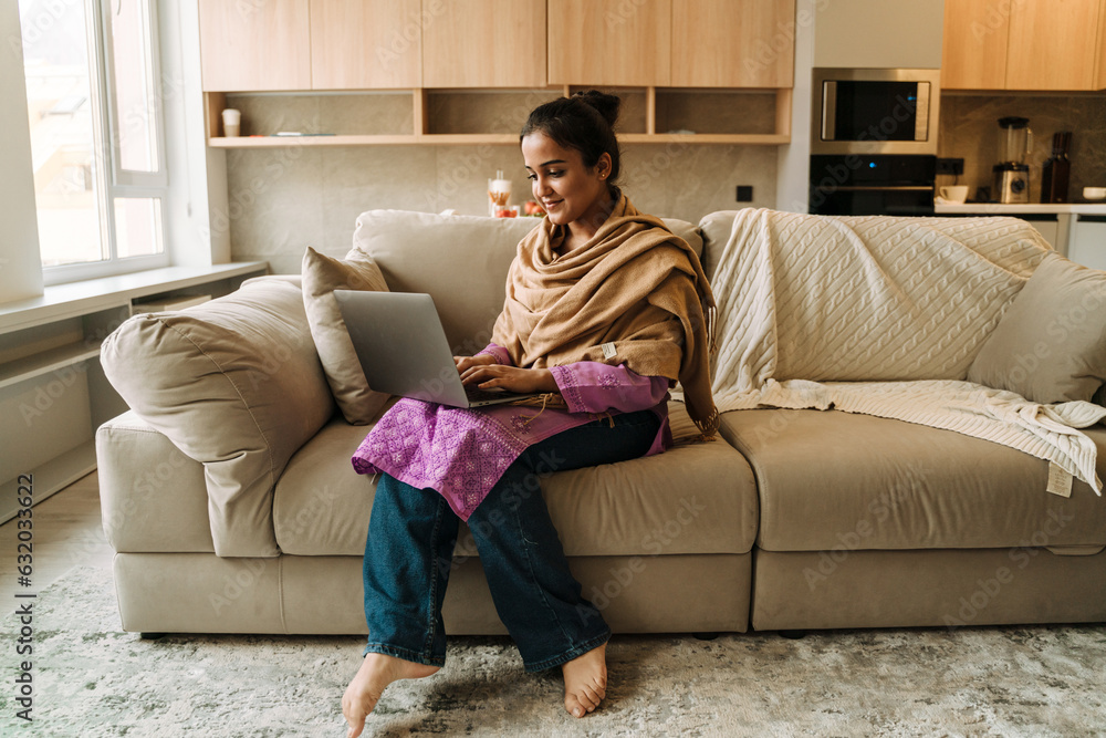 Young indian woman smiling and using laptop while sitting on couch Young indian woman smiling and using laptop while sitting on couch