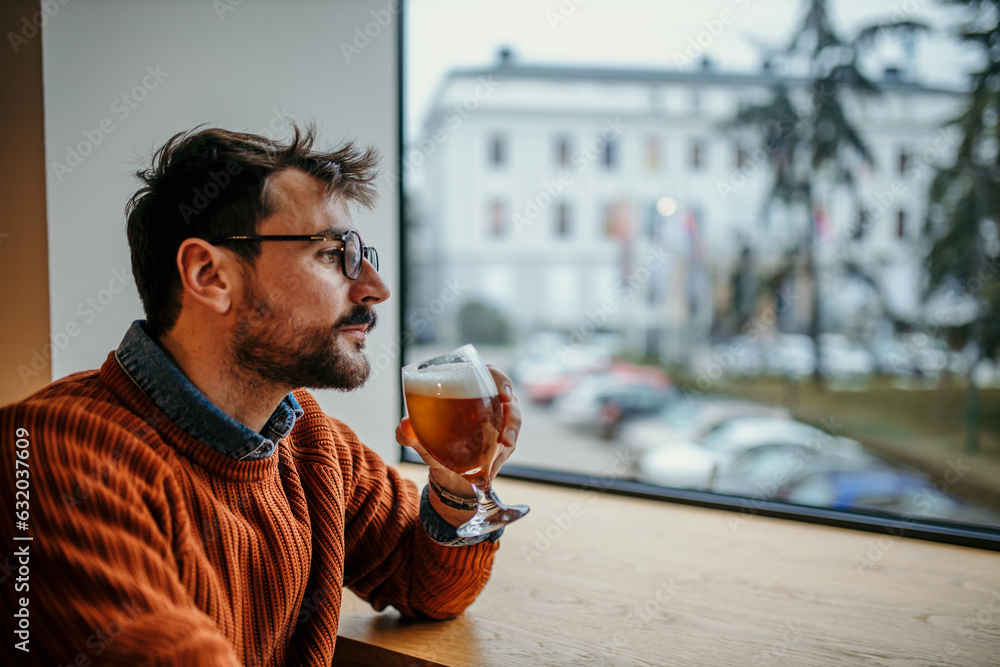 Fototapeta premium A smiling man sitting in the restaurant, wearing a smart casual outfit, using a phone, and drinking a beer.