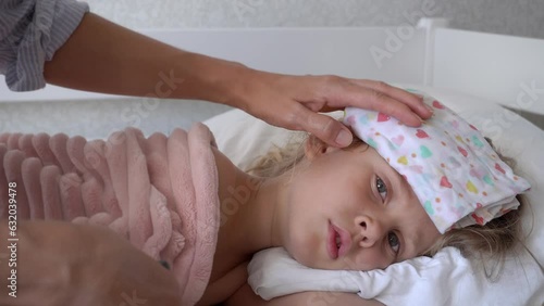 A sick girl lies in bed with a wet towel on her head. Mother holds a wet towel on the forehead of a sick girl. A child has a high temperature.