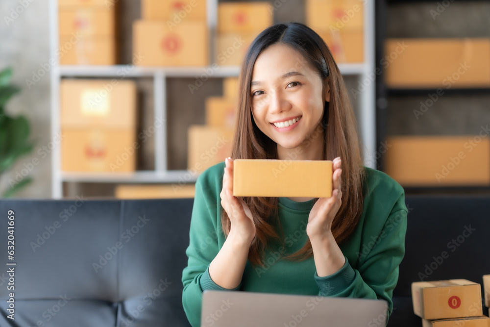 Smiling young Asian business owner woman prepare parcel box and check online orders of product for deliver to customer on laptop computer at home. Shopping Online concept. Startup small business SME. Smiling young Asian business owner woman prepare parcel box and check online orders of product for deliver to customer on laptop computer at home. Shopping Online concept. Startup small business SME.