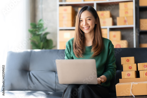 Smiling young Asian business owner woman prepare parcel box and check online orders of product for deliver to customer on laptop computer at home. Shopping Online concept. Startup small business SME. © Premreuthai