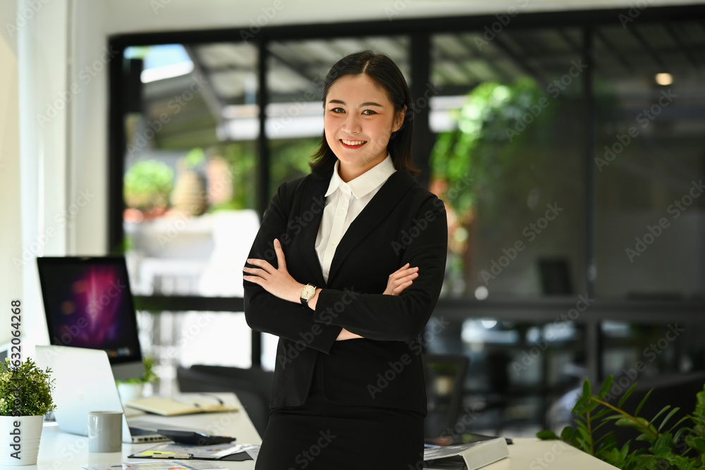 Portrait of confident millennial businesswoman in black suit standing with arms crossed and smiling at camera Portrait of confident millennial businesswoman in black suit standing with arms crossed and smiling at camera