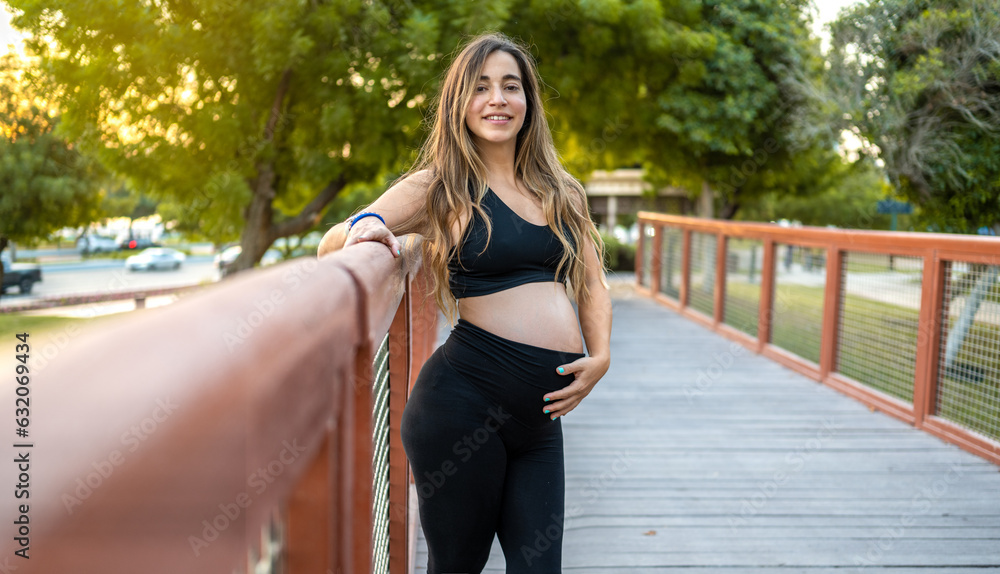 Smiling pregnant woman leaning over bridge fence, touching her belly and looking at camera outdoors.