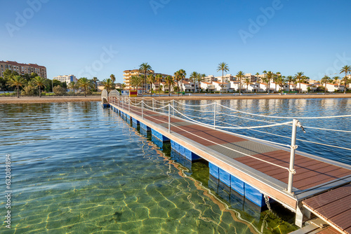 Iron bridge and wooden walkway over the crystalline beach of El Mar de Cristal, in the Mar Menor, Cartagena, Spain with the town in the background