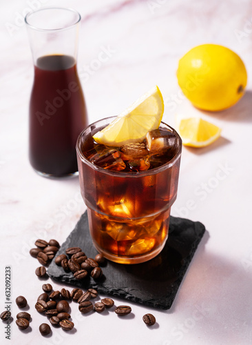 Cold brew coffee in a glass with ice and lemon on a light marble background with coffee beans and bottle. Concept summer craft refreshing homemade drink