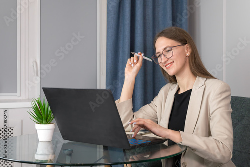 Fotografie Beautiful smiling young woman with a pen in her hands, wearing glasses and a jacket, is working at a laptop sitting at a table