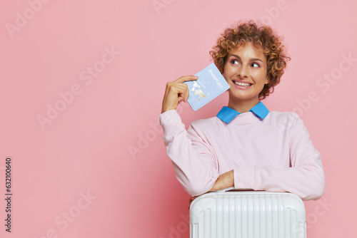 Curly cute girl wearing white sweatshot posing on pink background with passport in hand and white suitcase, happy tourist concept, copy space photo