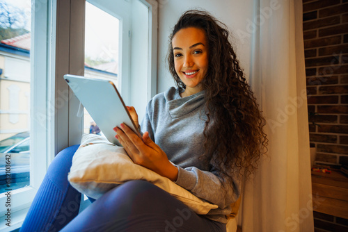 Young student sitting in a bay window, thinking about project ideas and making plans on her tablet