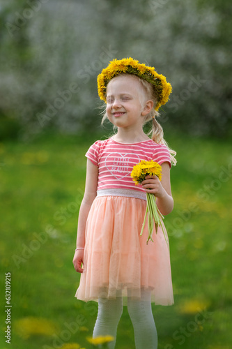 Happy blond girl with closed eyes wearing dandelion wreath outside. Child with bouquet of dandelions. Blurred background. Vertical frame photo