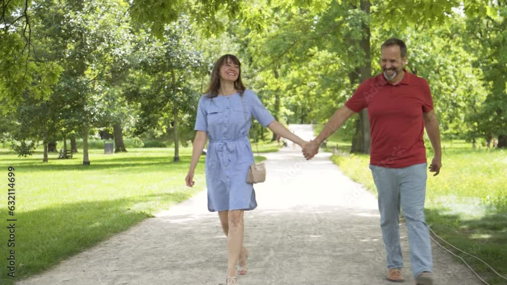 A middle-aged Caucasian couple holds hands and hugs as they walk down a pathway through a park on a sunny day A middle-aged Caucasian couple holds hands and hugs as they walk down a pathway through a park on a sunny day