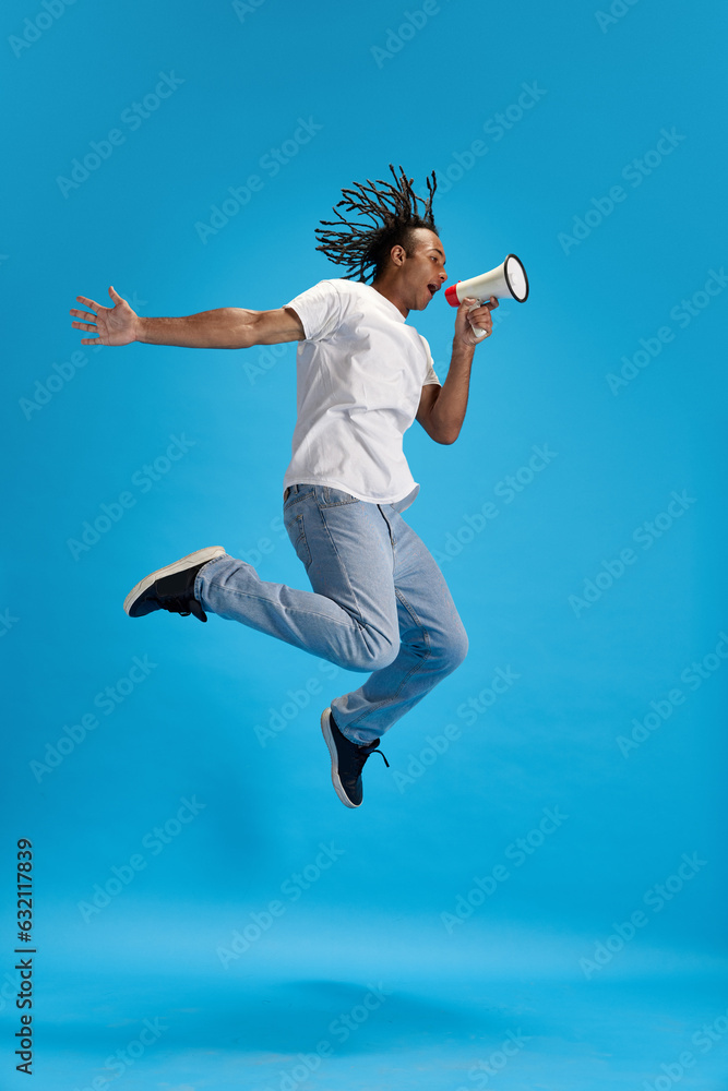 Announcement. News. Young guy in casual clothes jumping and shouting in megaphone against blue studio background. Concept of youth, human emotions, lifestyle, fashion, sales, ad
