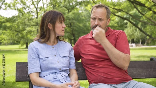 A middle-aged Caucasian couple talks about something and acts upset as they sit on a bench in a park