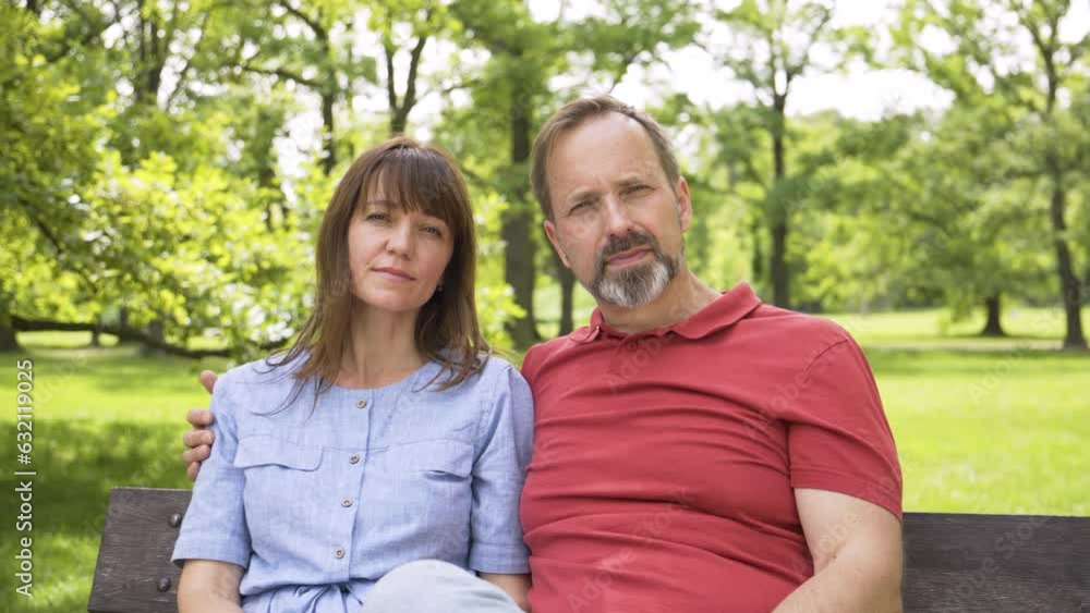 A middle-aged Caucasian couple looks seriously at the camera as they sit on a bench in a park on a sunny day - ends in closeup A middle-aged Caucasian couple looks seriously at the camera as they sit on a bench in a park on a sunny day - ends in closeup