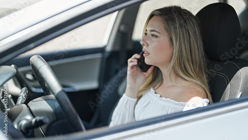 Young blonde woman talking on smartphone sitting on car at street