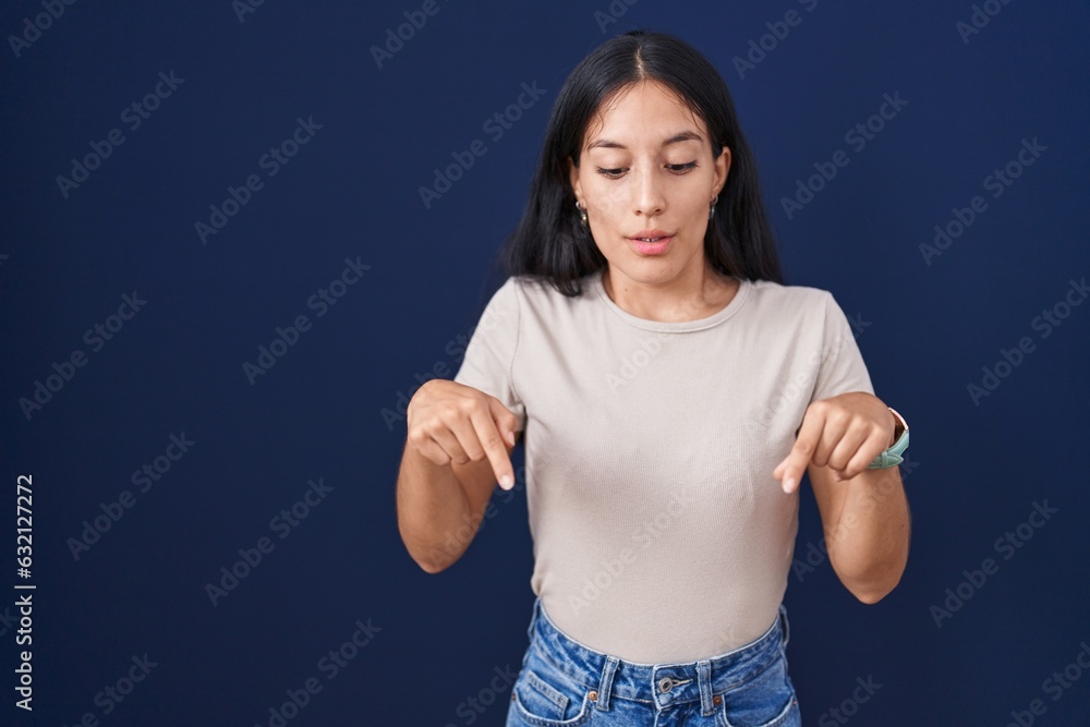 Young hispanic woman standing over blue background pointing down with fingers showing advertisement, surprised face and open mouth Young hispanic woman standing over blue background pointing down with fingers showing advertisement, surprised face and open mouth