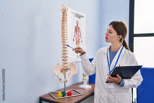 Young blonde woman pysiotherapist pointing to anatomical model of vertebral column holding clipboard at rehab clinic