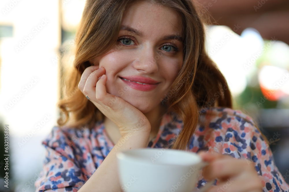 A cheerful young woman enjoying a hot coffee in a summer morning. She is holding a white cup and smiling while sitting at an outdoor cafe with green plants. A cheerful young woman enjoying a hot coffee in a summer morning. She is holding a white cup and smiling while sitting at an outdoor cafe with green plants.