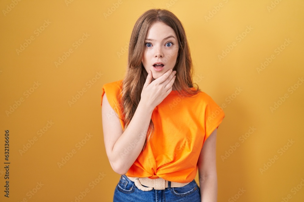 Caucasian woman standing over yellow background looking fascinated with disbelief, surprise and amazed expression with hands on chin Caucasian woman standing over yellow background looking fascinated with disbelief, surprise and amazed expression with hands on chin