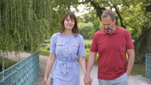 A middle-aged Caucasian couple walks across a bridge in a park on a sunny day, talks and smiles - front view