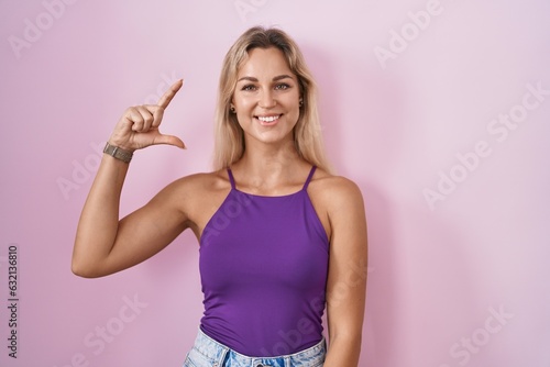 Young blonde woman standing over pink background smiling and confident gesturing with hand doing small size sign with fingers looking and the camera. measure concept. © Krakenimages.com
