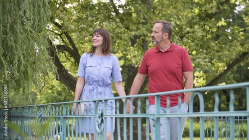 A middle-aged Caucasian couple on a walk stops on a bridge at the railing and looks around with smiles in a park on a sunny day