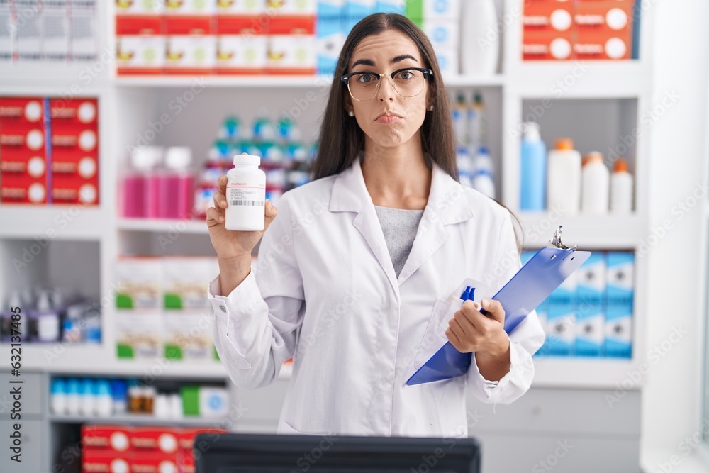 Young brunette woman working at pharmacy drugstore holding pills depressed and worry for distress, crying angry and afraid. sad expression.