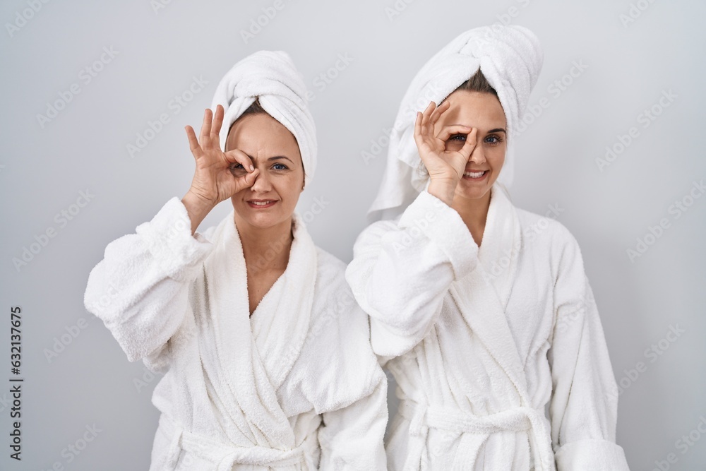 Middle age woman and daughter wearing white bathrobe and towel doing ok gesture with hand smiling, eye looking through fingers with happy face. Middle age woman and daughter wearing white bathrobe and towel doing ok gesture with hand smiling, eye looking through fingers with happy face.
