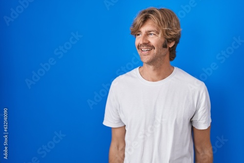 Middle age man standing over blue background looking away to side with smile on face, natural expression. laughing confident.