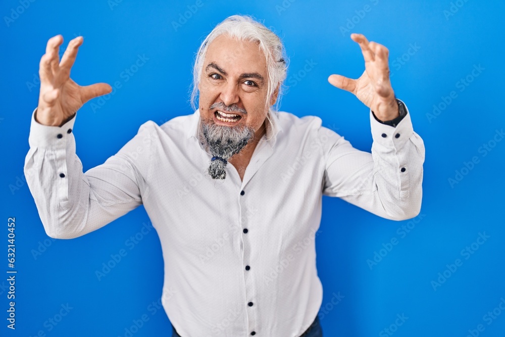Middle age man with grey hair standing over blue background shouting frustrated with rage, hands trying to strangle, yelling mad Middle age man with grey hair standing over blue background shouting frustrated with rage, hands trying to strangle, yelling mad