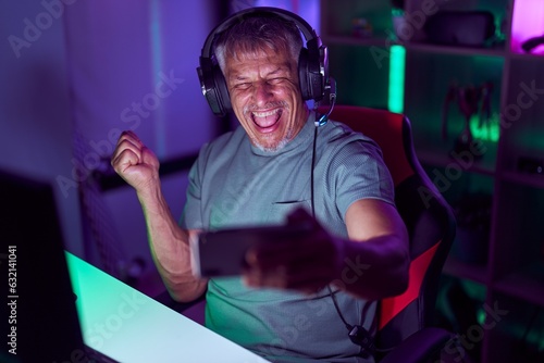 Hispanic man with grey hair playing video games with smartphone screaming proud, celebrating victory and success very excited with raised arm © Krakenimages.com