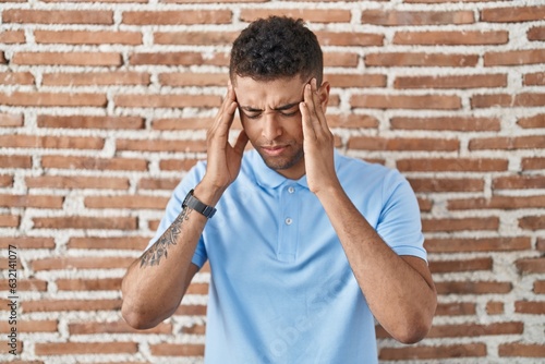 Brazilian young man standing over brick wall with hand on head, headache because stress. suffering migraine.