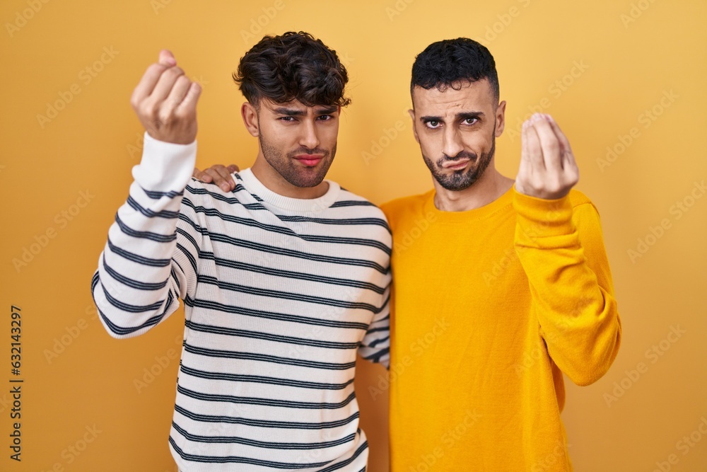 Young hispanic gay couple standing over yellow background doing italian gesture with hand and fingers confident expression Young hispanic gay couple standing over yellow background doing italian gesture with hand and fingers confident expression