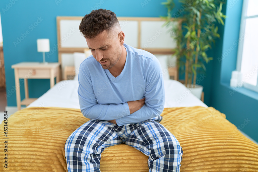 Young hispanic man suffering for stomach ache sitting on bed at bedroom Young hispanic man suffering for stomach ache sitting on bed at bedroom