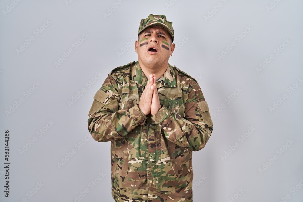 Hispanic young man wearing camouflage army uniform begging and praying with hands together with hope expression on face very emotional and worried. begging. Hispanic young man wearing camouflage army uniform begging and praying with hands together with hope expression on face very emotional and worried. begging.