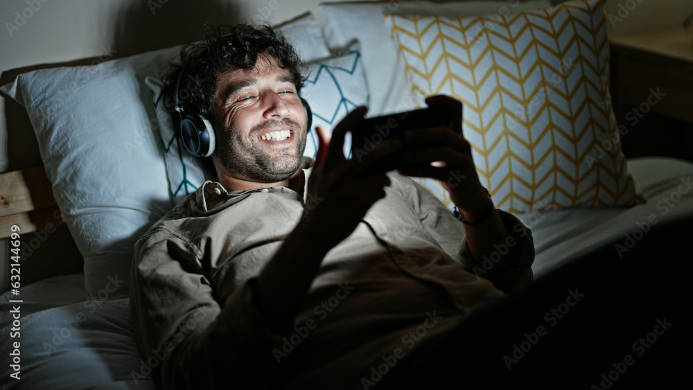 Fototapeta premium Young hispanic man watching video on smartphone lying on bed at bedroom
