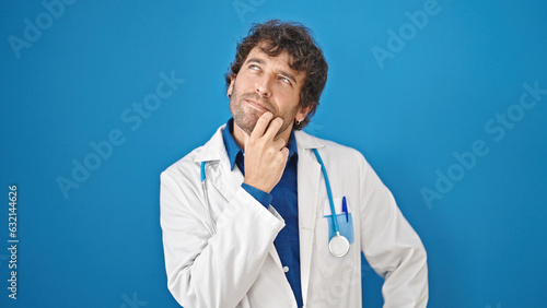 Young hispanic man doctor standing with doubt expression thinking over isolated blue background
