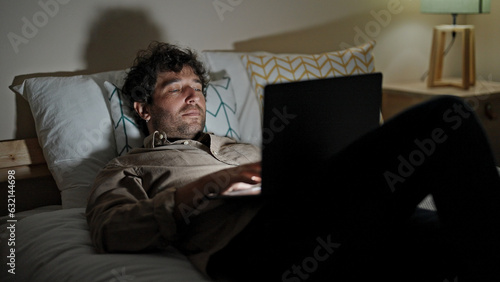 Young hispanic man using laptop lying on bed working at bedroom