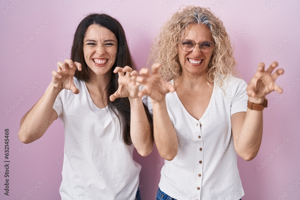 Mother and daughter standing together over pink background smiling funny doing claw gesture as cat, aggressive and sexy expression Mother and daughter standing together over pink background smiling funny doing claw gesture as cat, aggressive and sexy expression