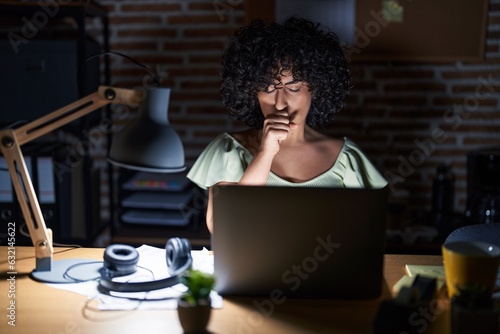 Young brunette woman with curly hair working at the office at night feeling unwell and coughing as symptom for cold or bronchitis. health care concept.