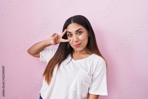 Young arab woman standing over pink background doing peace symbol with fingers over face, smiling cheerful showing victory