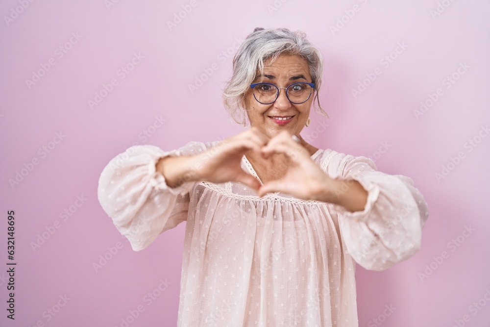 Middle age woman with grey hair standing over pink background smiling in love doing heart symbol shape with hands. romantic concept. Middle age woman with grey hair standing over pink background smiling in love doing heart symbol shape with hands. romantic concept.