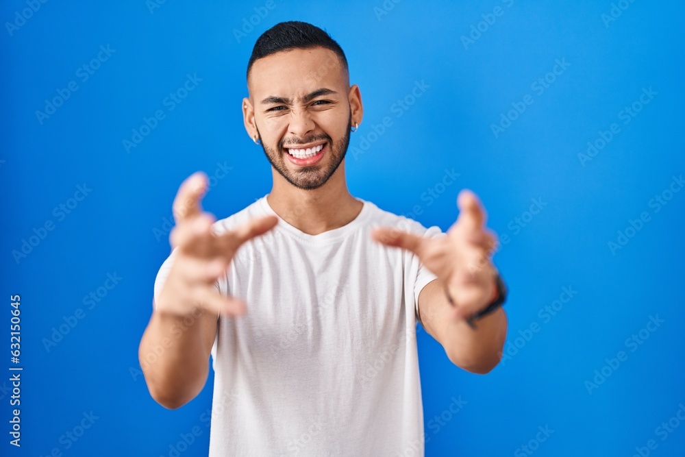 Young hispanic man standing over blue background shouting frustrated with rage, hands trying to strangle, yelling mad Young hispanic man standing over blue background shouting frustrated with rage, hands trying to strangle, yelling mad